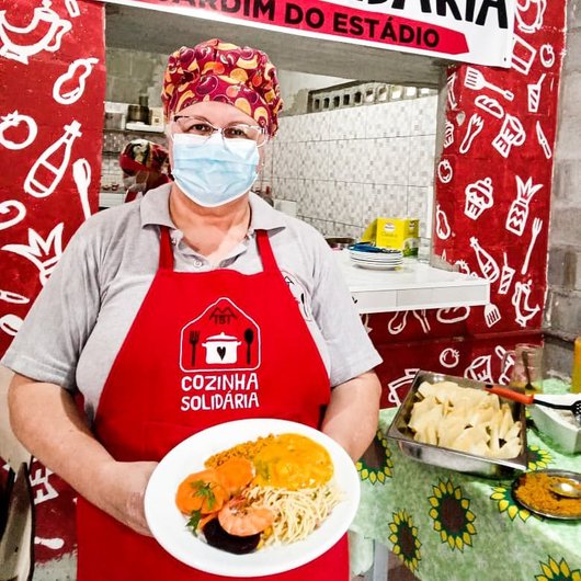 Foto de uma senhora com avental vermelho onde se lê 'cozinha solidária', segurando um prato com comida