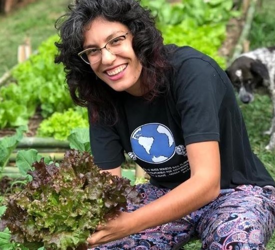 Foto de uma moça sorridente com calça colorida e camiseta preta agachada em meio a uma plantação, segurando um pé de alface